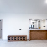 Empty bedroom in a newly renovated high-rise apartment, featuring light gray large-format porcelain floor tiles, crisp white walls, recessed ceiling lighting, a large black-framed corner window providing abundant natural light and direct views of surrounding urban high-rise buildings, a partially open light wood door leading to an en-suite or hallway, and a wall-mounted air-conditioning unit visible through the doorway.